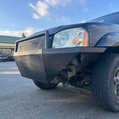 Modified truck front with angular black bumper and rugged tire, parked outdoors.