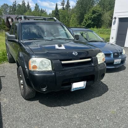 Black Nissan SUV parked on gravel near a blue car in a green outdoor setting.