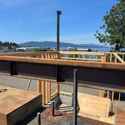 Construction site view with steel beam, framing, and scenic mountains and water in background.