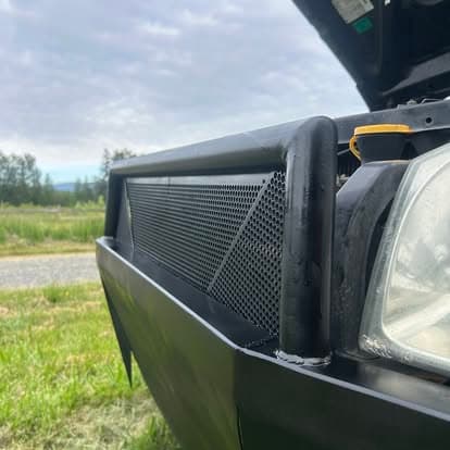 Custom truck front grille with mesh design and auxiliary light, set against a rural backdrop.